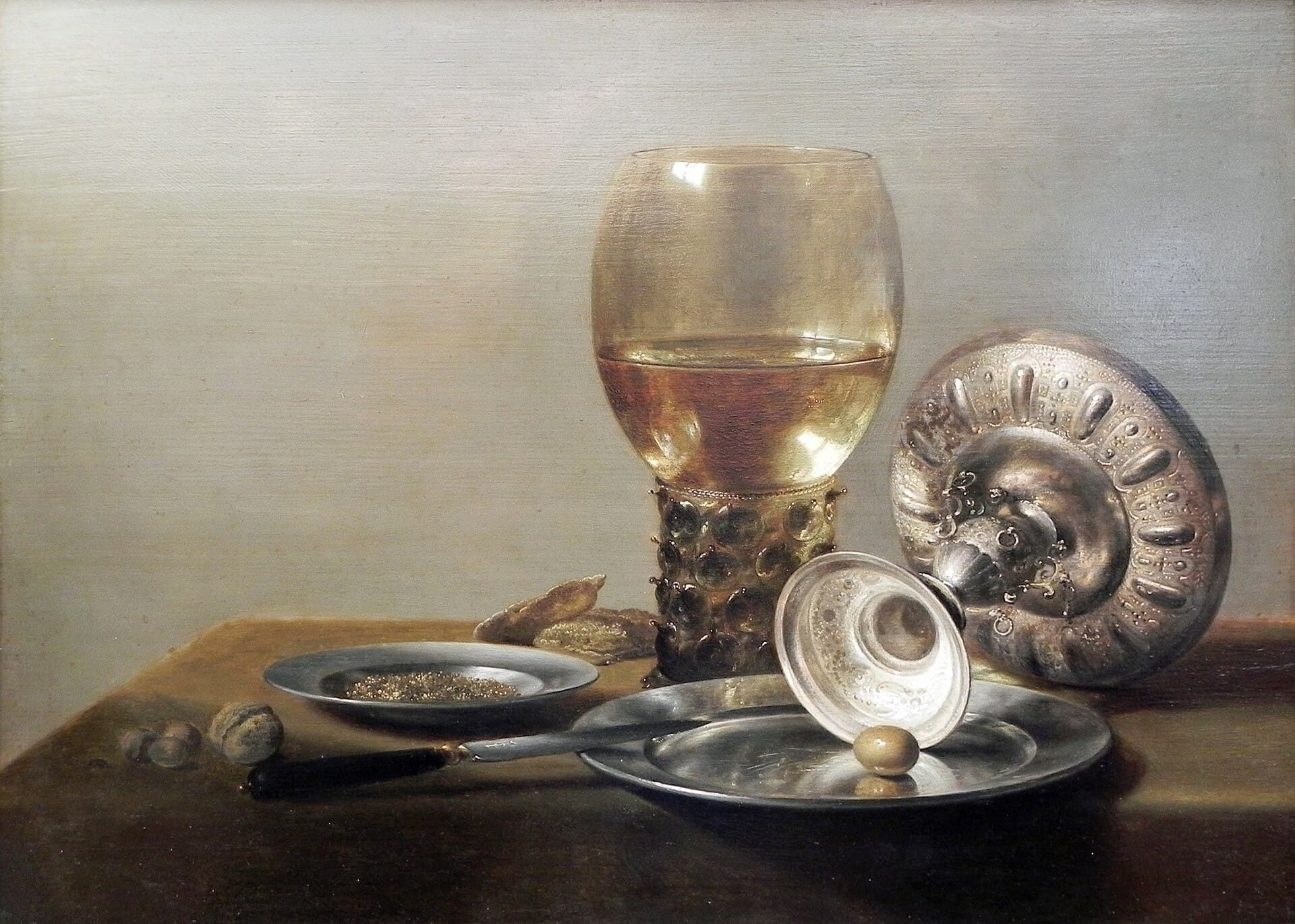 A Dutch still-life painting of a tall roemer glass of white wine beside an overturned silver tazza, a pewter plate and walnuts on a table.